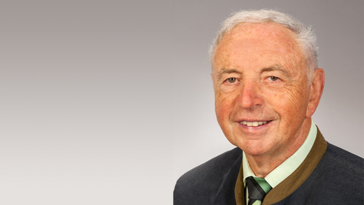 Ein älterer Mann mit grauen Haaren lautet die Beschreibung dieses Bildes. Er lächelt freundlich und trägt ein dunkles Sportjackett über einem schweigens Hemd mit Krawatte.