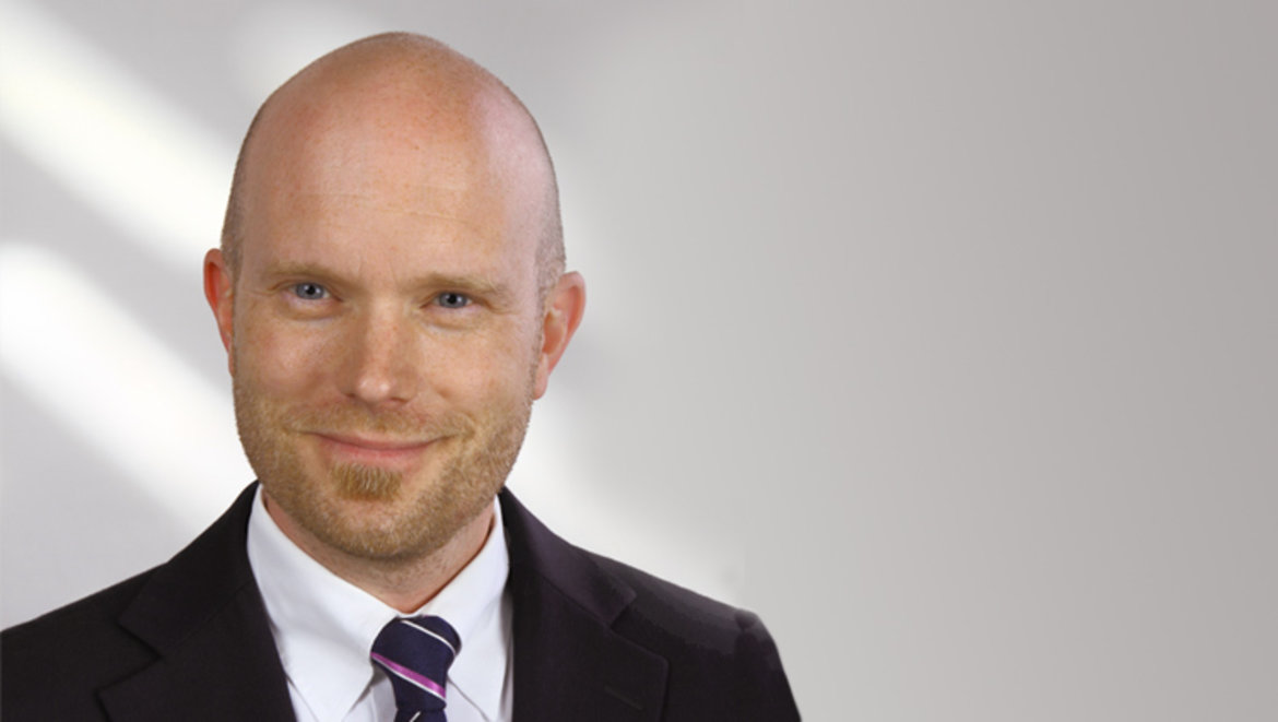 Ein lächelnder Mensch mit glattrasiertem Haupt, weißem Hemd, violett gestreifter Krawatte und dunklem Jackett.

 prennent den Raum ein;
 die Aufnahme wirkt professionell.