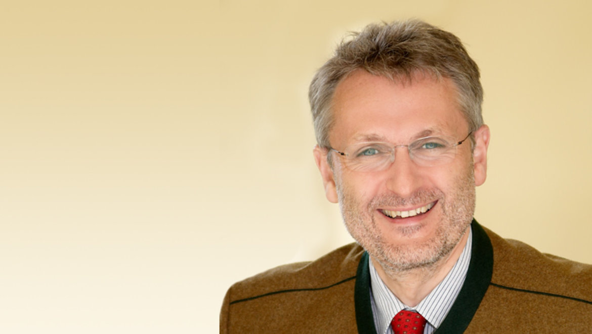 Ein lächelnder Mann mit grauen Haaren und Brille, der einen grünbraunen Blazer und eine rote Krawatte trägt. Professionell und freundlicher Ausdruck.